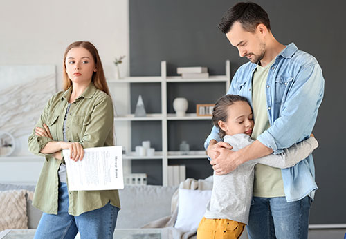 Father hugging his daughter while the mother stands with crossed arms over child custody papers