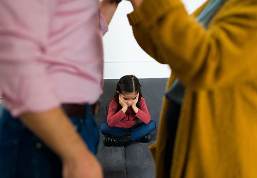 Upset child sitting alone while her arguing parents stand in the background, needing custody modification