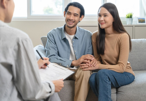 A smiling couple holding hands, sitting on a couch across from a person reviewing papers.